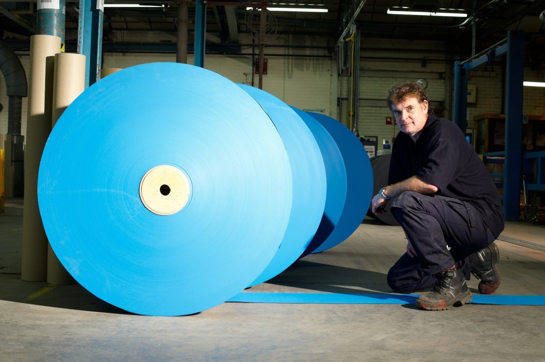 Man crouching beside a large blue roll of paper in an industrial factory setting.