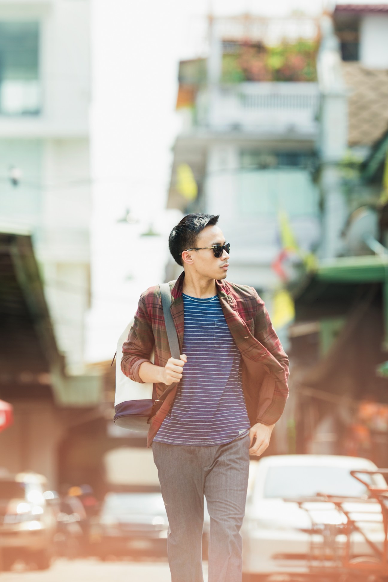 Man wearing sunglasses, burgundy jacket, striped t-shirt and jeans walks through Bangkok alleyway.