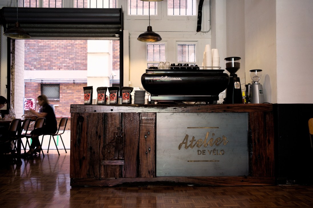 Espresso bar counter at Atelier de Vélo with black La Marzocco machine and wooden cabinetry.