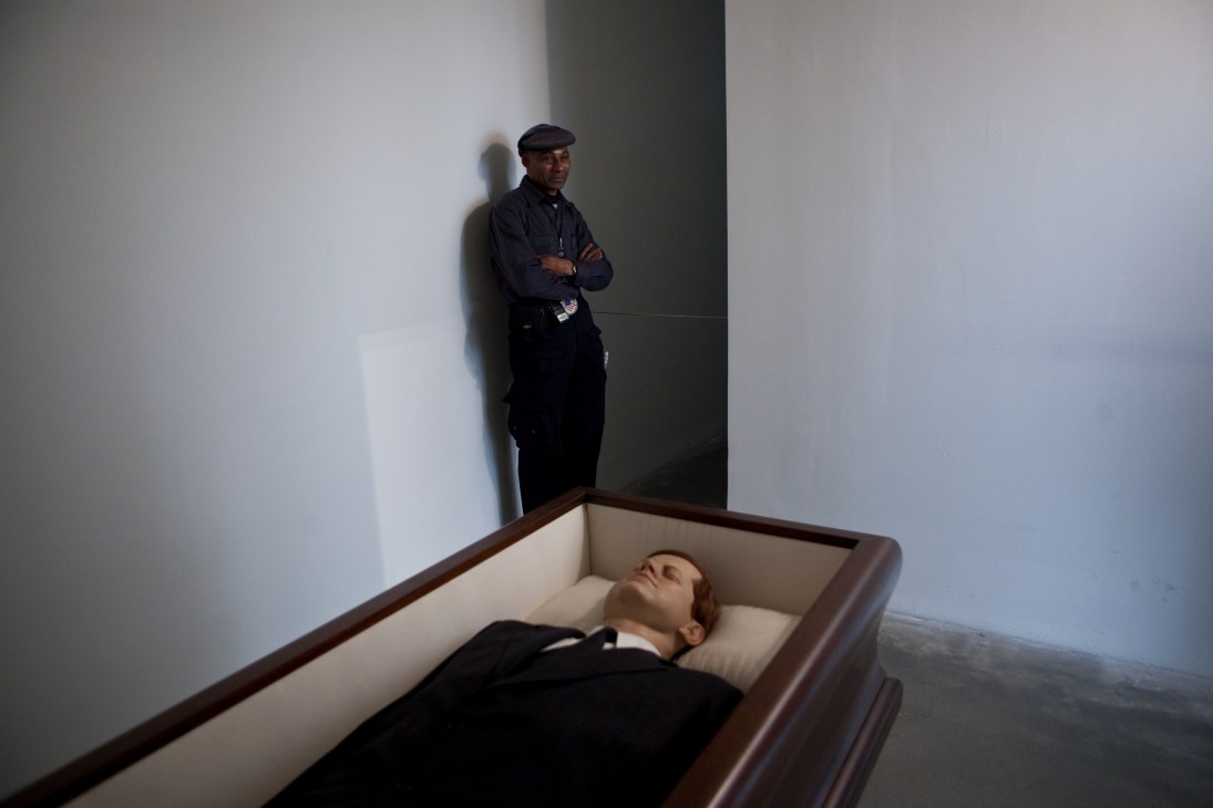 Maurizio Cattelan's 'Now' (2004), an artwork featuring a figure lying in a coffin with a man standing beside it.