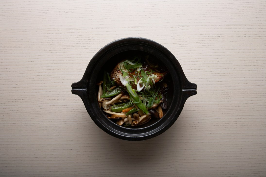 Miso Chilean seabass toban yaki served in a black ceramic bowl with green onions and mushrooms on a wooden surface.