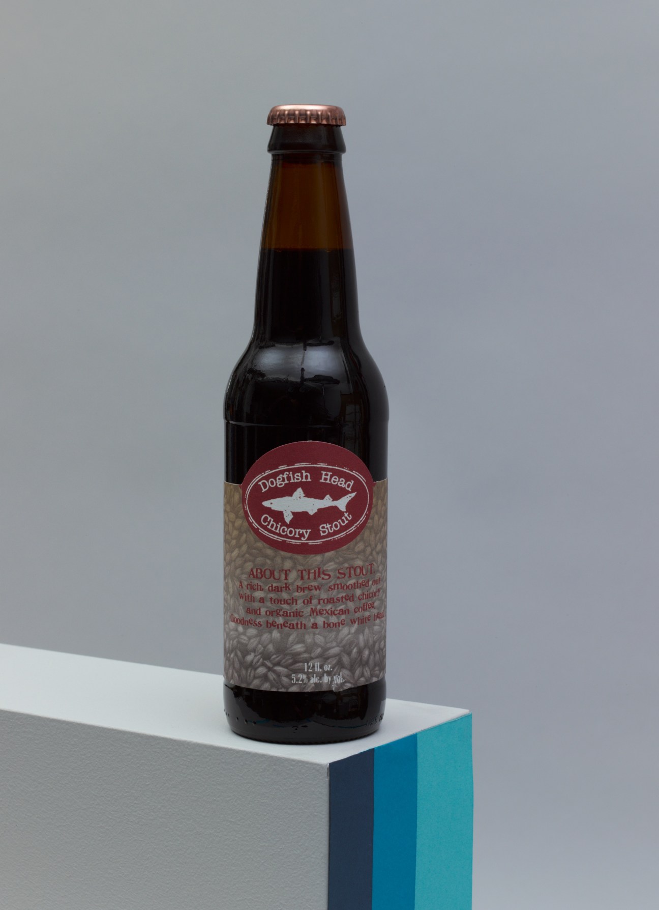 Dogfish Head Chicory Stout beer bottle with red label and copper cap on a geometric blue and white base.