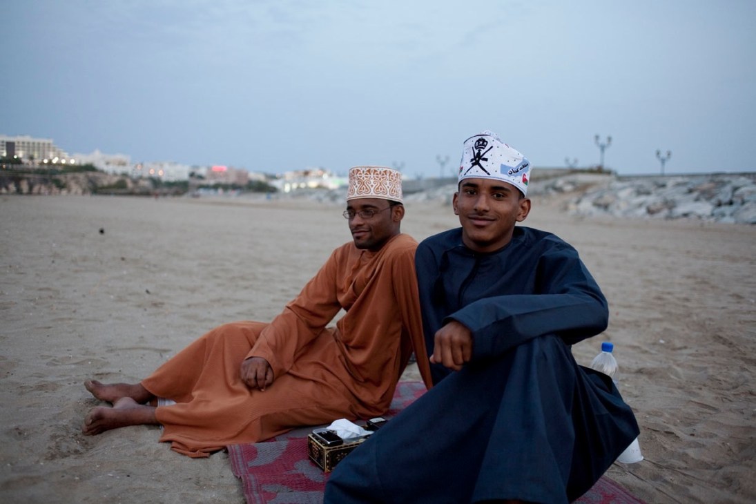Two young men sit on a beach in Muscat wearing traditional Omani clothing and caps.