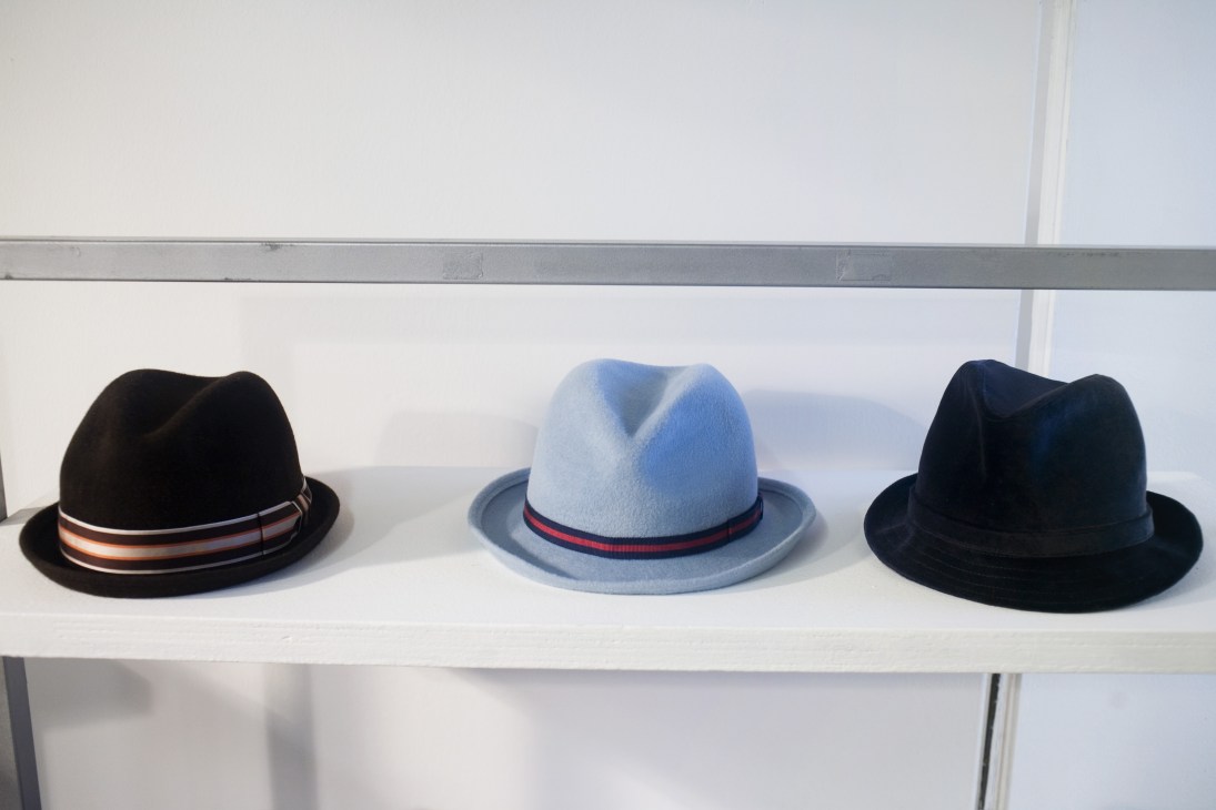 Three Anthony Peto fedora hats displayed on a white shelf: black with striped band, light blue with navy-red band, and navy blue.