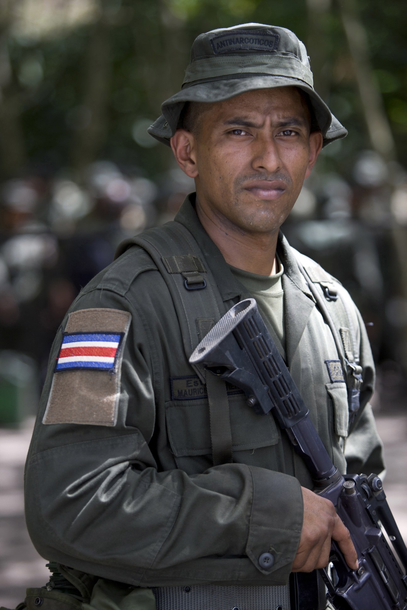 A Colombian anti-narcotics police officer wearing a bucket hat and tactical gear with a Costa Rican flag patch holds a rifle.