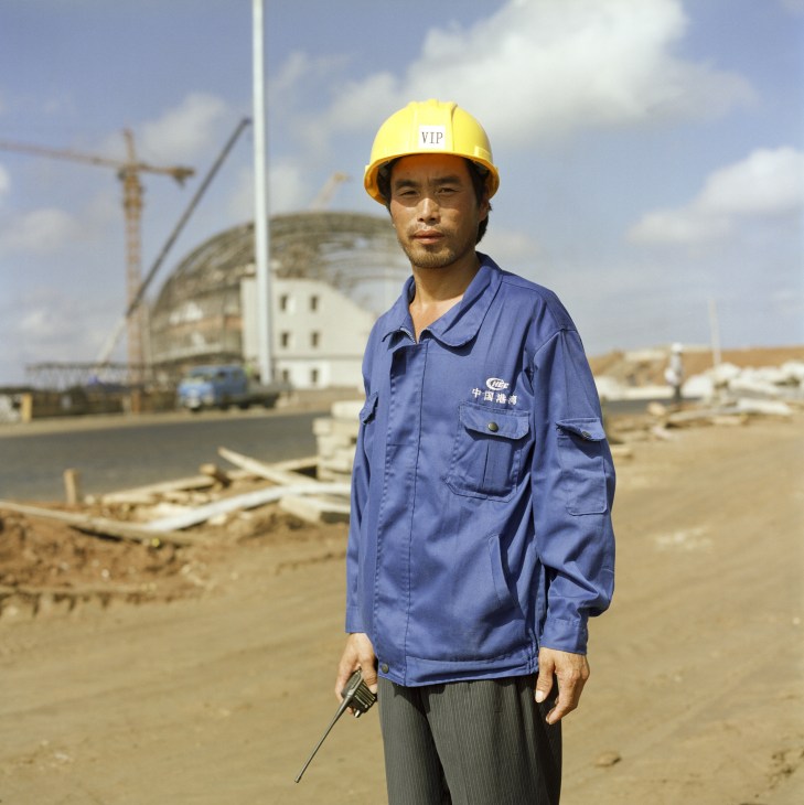 A Chinese worker in blue jumpsuit and yellow VIP helmet stands at Magampura Port construction site with stadium structure visible behind.