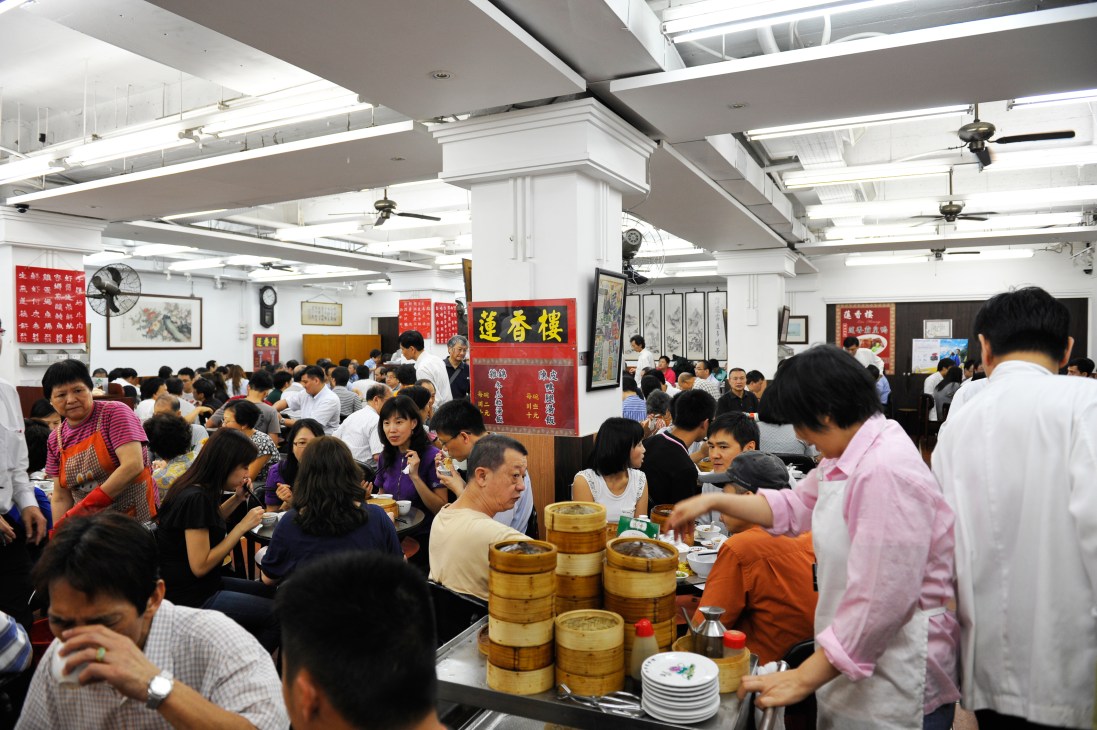 Crowded dim sum restaurant interior with diners at tables and bamboo steamer baskets.