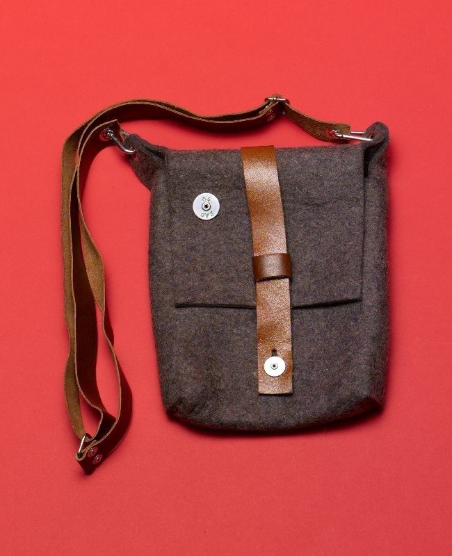 Carga felt and leather shoulder bag with brown leather strap and decorative ribbon closure on red background.