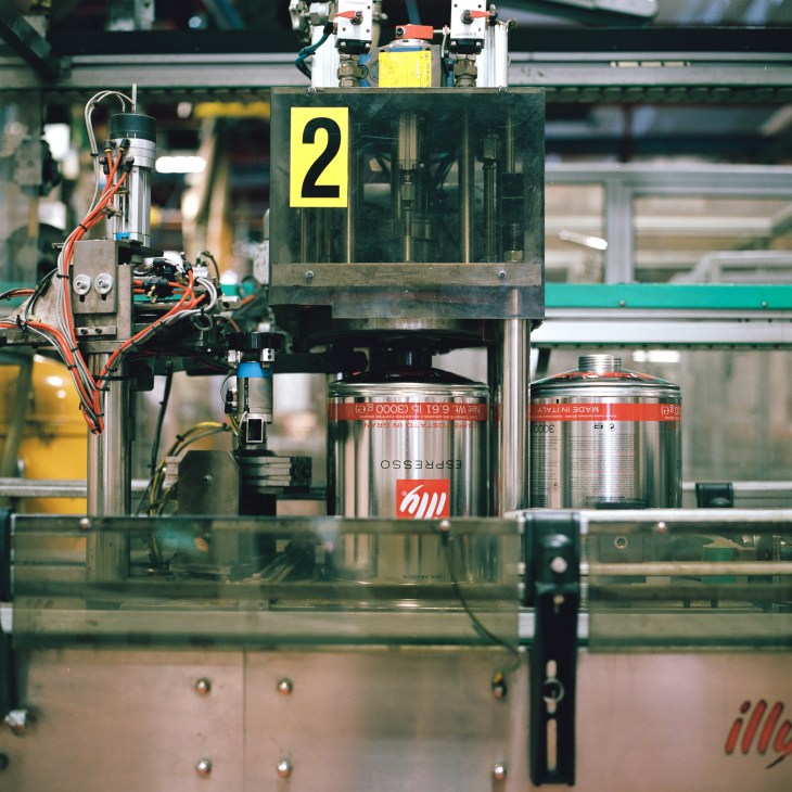 Illy coffee tins with red stripes move along an automated production line with mechanical equipment.