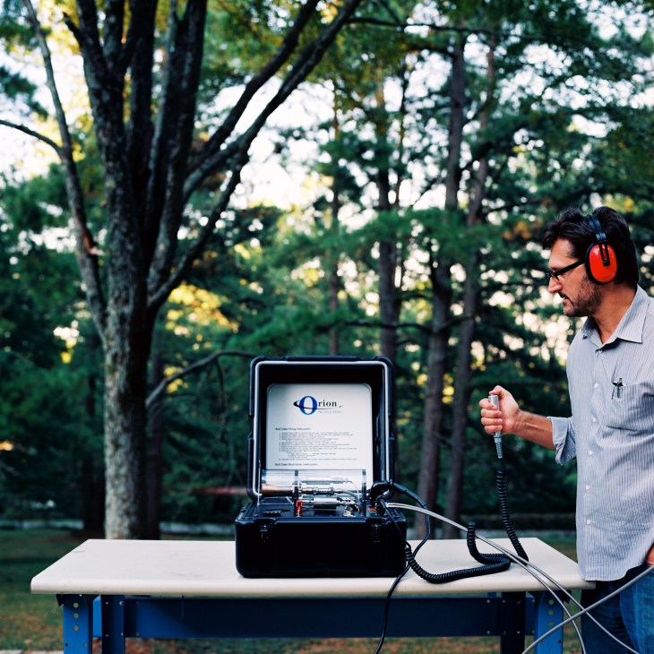 Tim Pickens wearing red headphones demonstrates his portable Orion satellite transmitter device outdoors under tall trees.