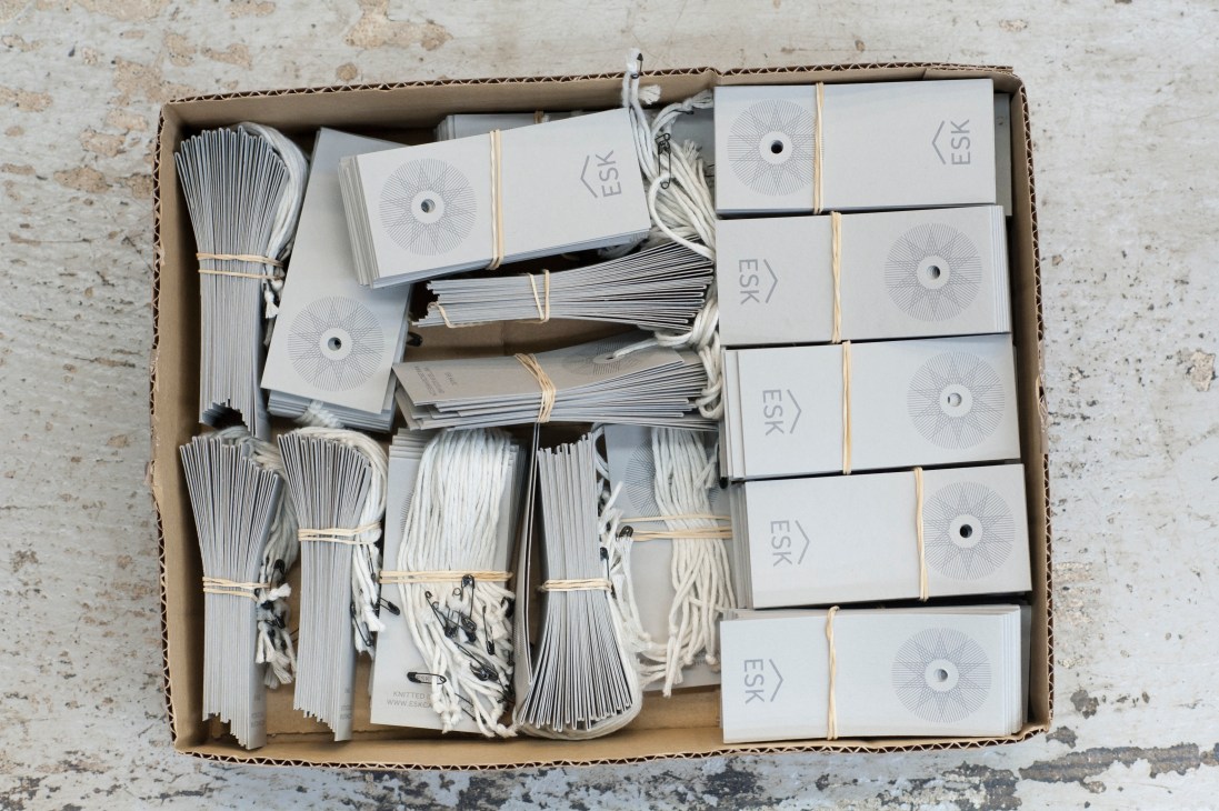Cardboard box containing white packaged Esk knitwear products with the logo and cream-colored ties, viewed from above.
