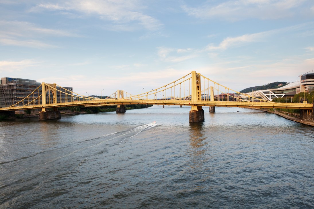 A bright yellow suspension bridge spans a river with modern buildings and hills visible in the background.