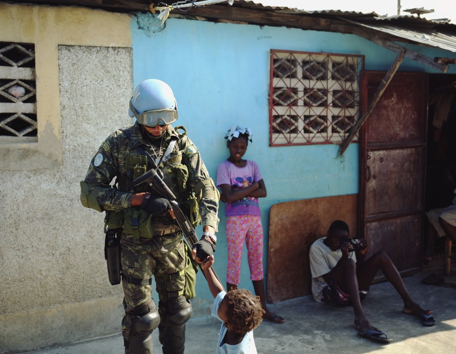 A Brazilian peacekeeper in full protective gear interacts with local children in a Haitian slum neighborhood.