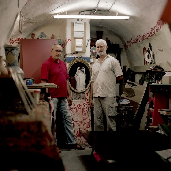 Artisan Mostafa Bouelkak in red shirt and casbah historian Hassan Smaallah stand in a narrow workshop with arched ceiling decorated with red patterns and ornaments.