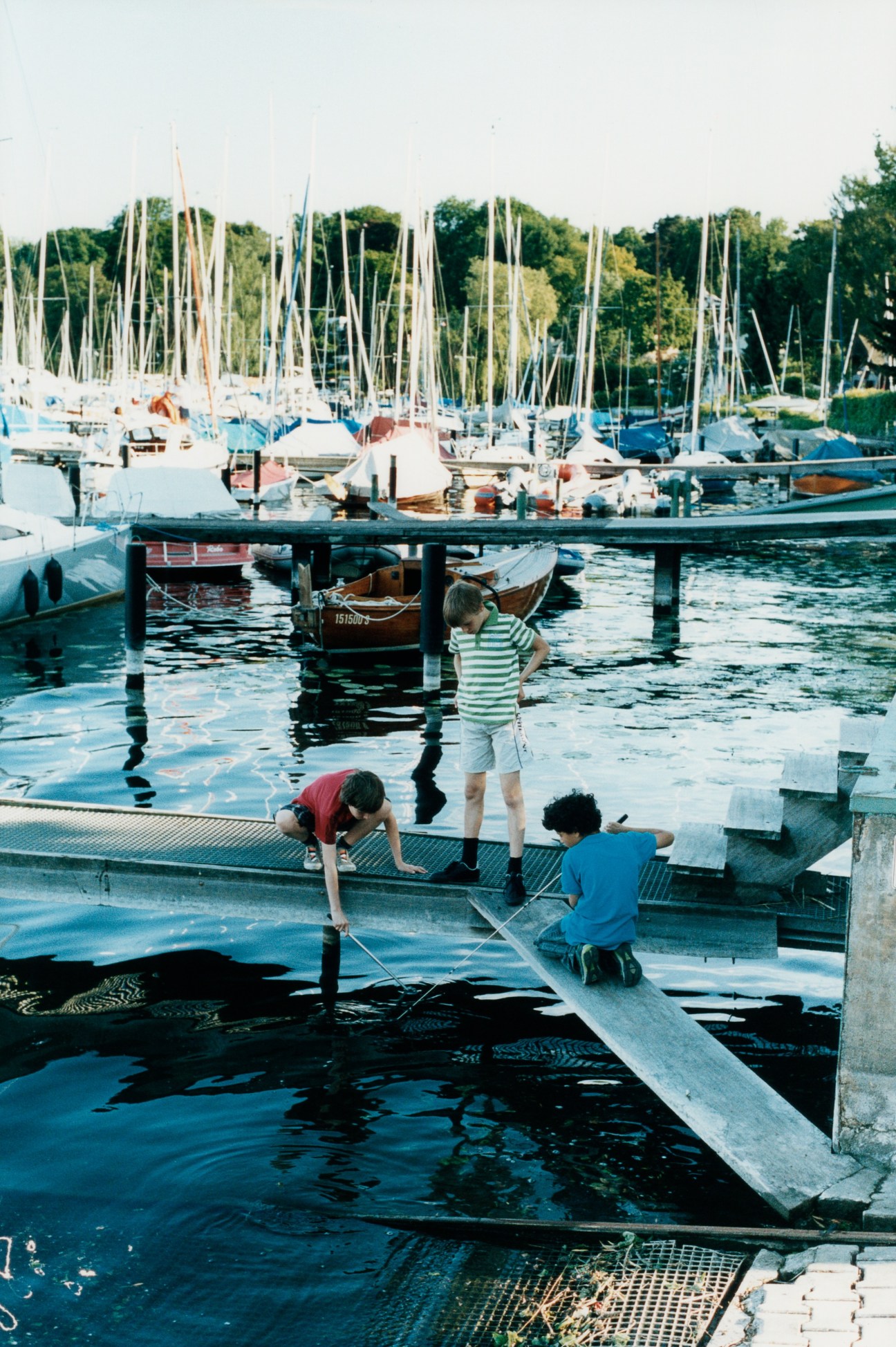 Children playing on a wooden dock at a lakeside sailing club with moored boats and trees in the background.