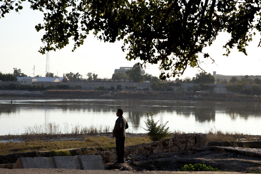 A person stands on the banks of the Tigris River in Baghdad's Abu Nawaz district, overlooking the water and buildings across the riverbank.