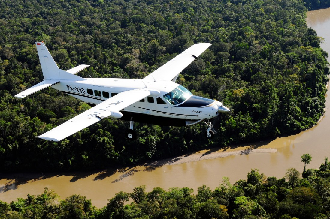 A white Cessna Caravan aircraft flies over dense jungle with a brown river visible below.