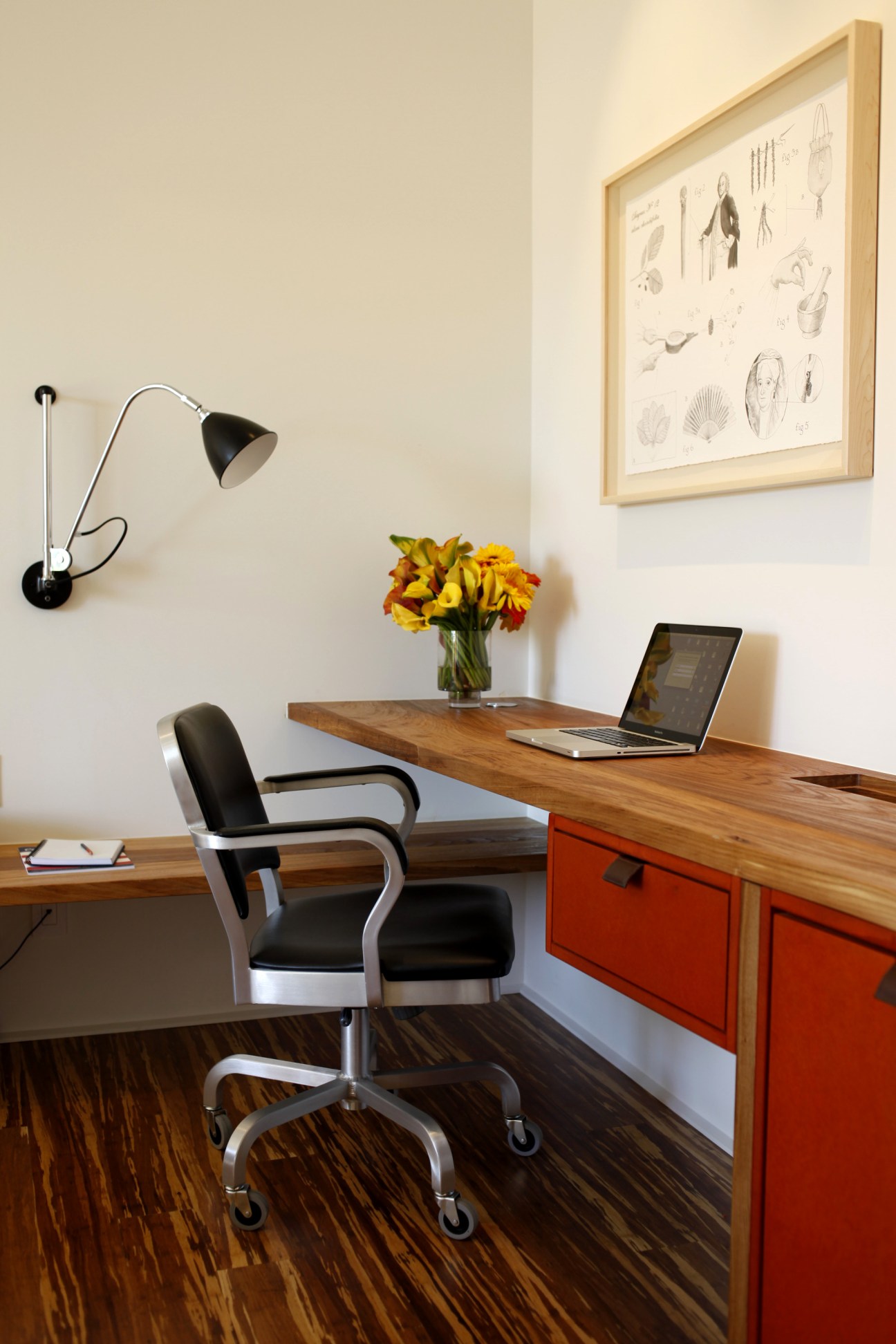 Modern hotel room workspace with wooden desk, red drawers, black chair, desk lamp and framed artwork on wall.