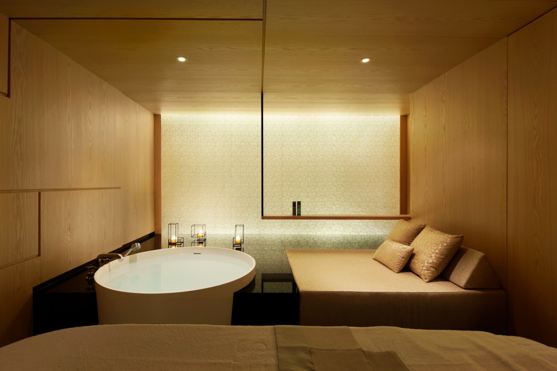 Minimalist spa treatment room with wooden walls, circular white bathtub, illuminated mirror, and cushioned seating area.