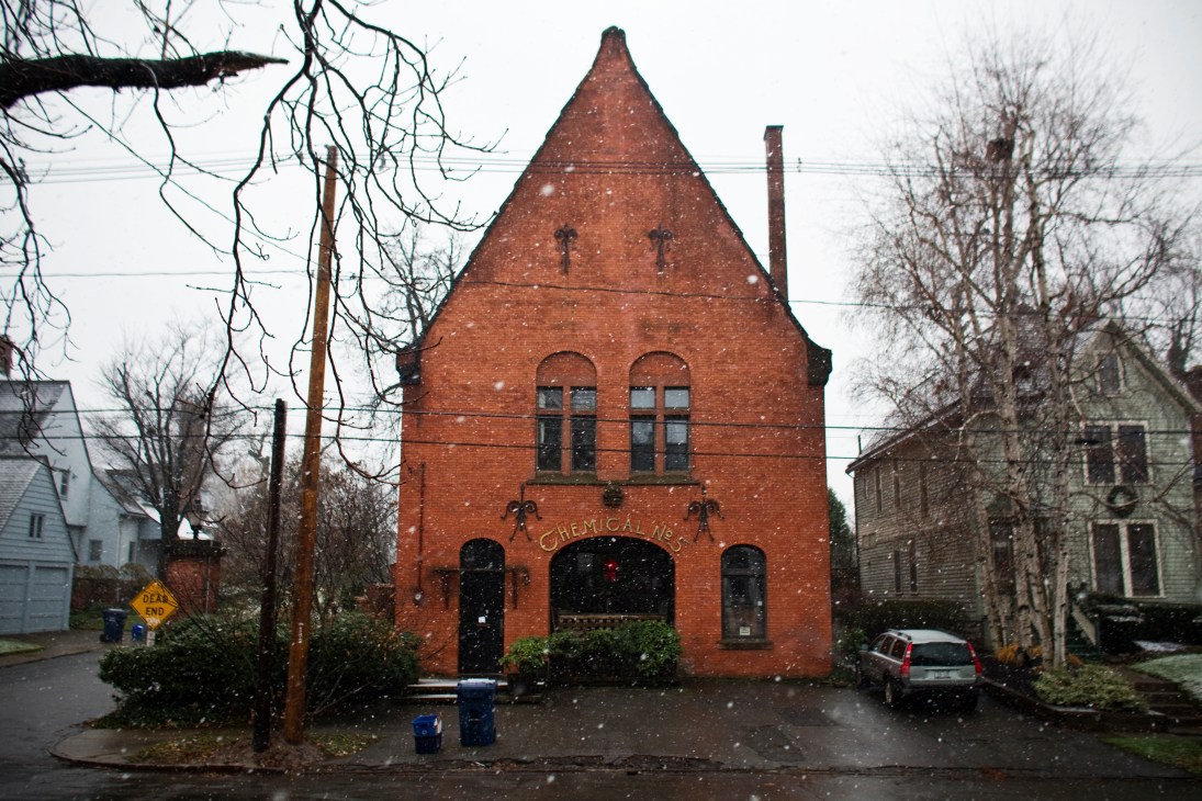 A red brick fire station building with peaked roof and arched windows converted into a residential home, surrounded by bare trees.