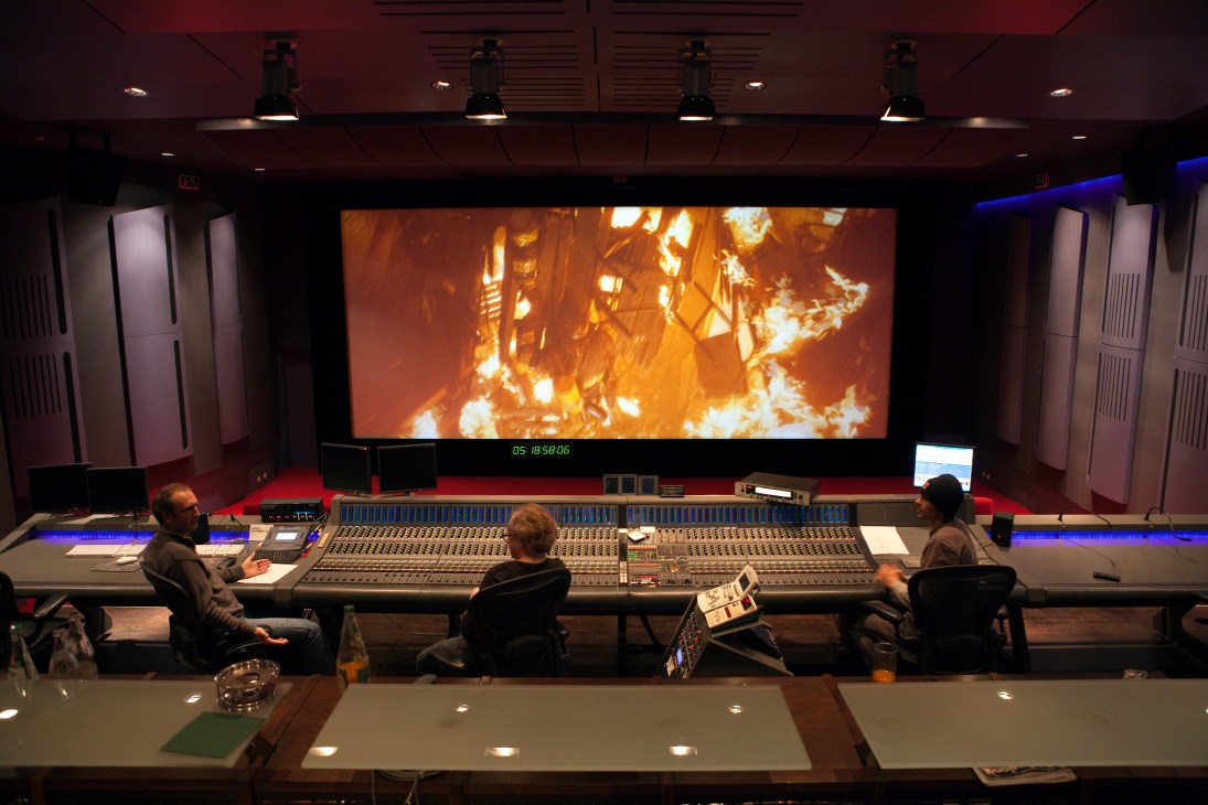 Three people work at a large mixing console in a professional dubbing studio with a cinema screen displaying fire footage.
