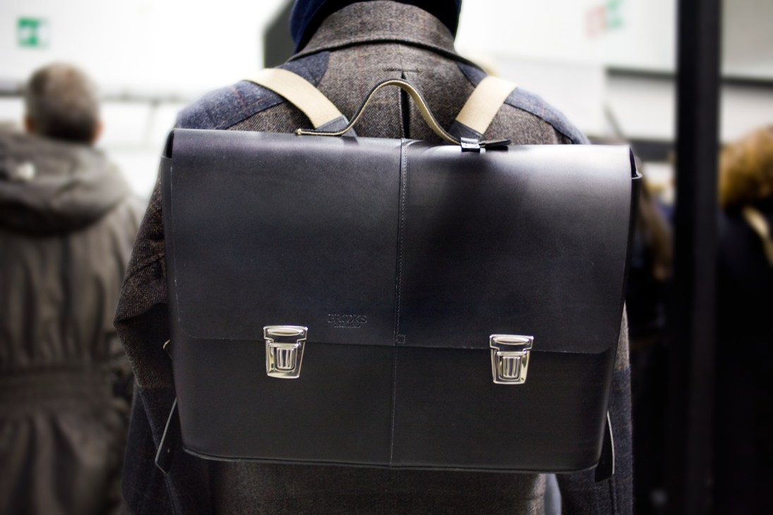 Black leather messenger bag with metal clasps worn on a person's back at a fashion event.