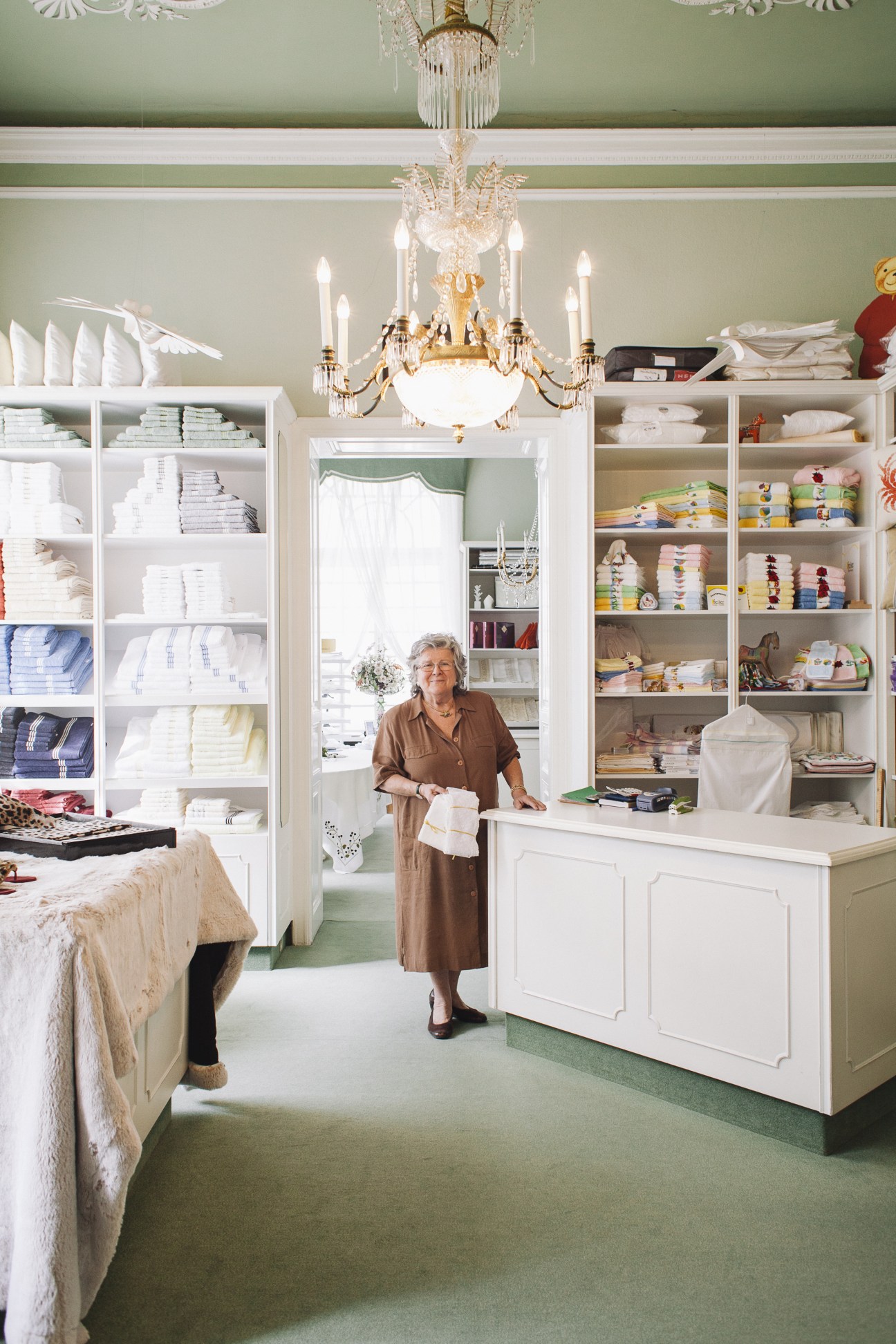 Interior of Zur Schwäbischen Jungfrau homeware shop with woman holding linens amid white shelving and chandelier.