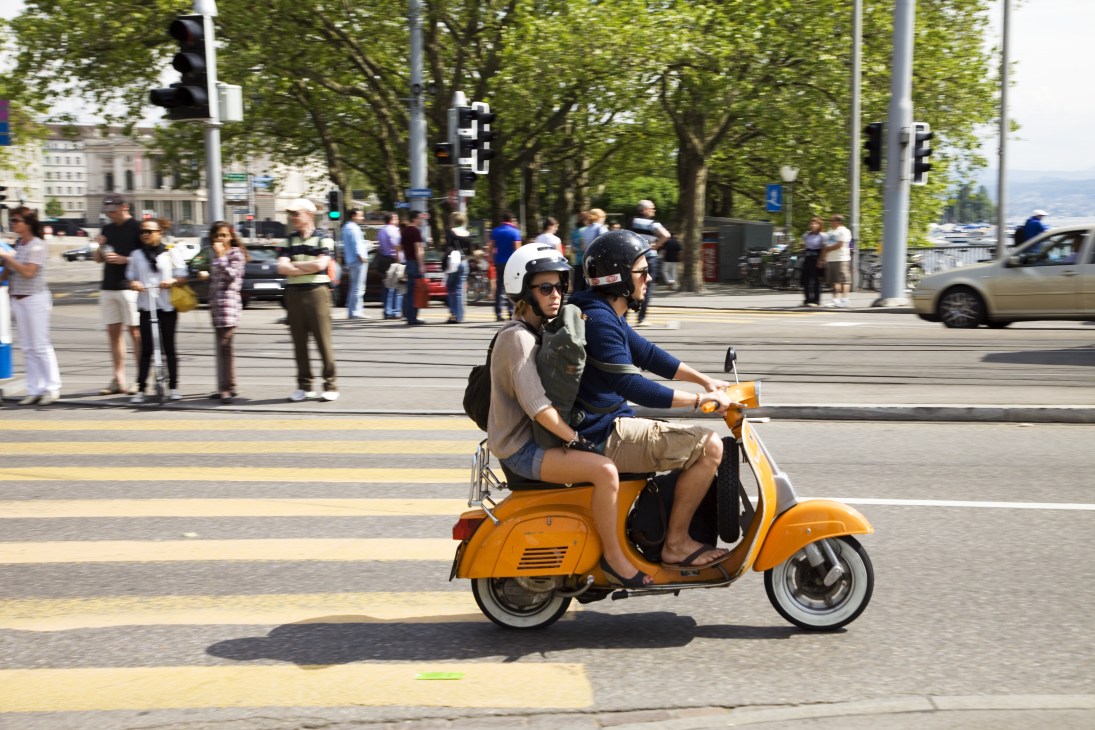 Two people wearing helmets ride a yellow Vespa scooter across a city street lined with trees and pedestrians.