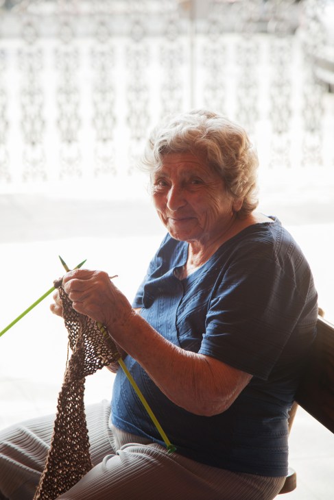 Elderly woman in blue shirt working on knitting with yarn and needles.
