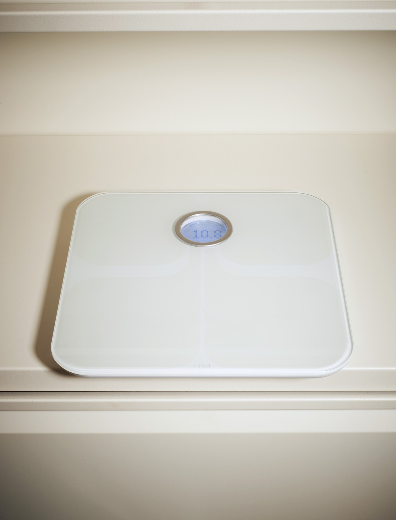 White Fitbit Smart Scale with blue digital display showing weight reading on beige shelf.