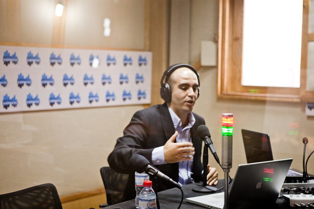 Radio show host Zied Mhirsi speaking into a microphone at a broadcasting studio desk with laptops and headphones.