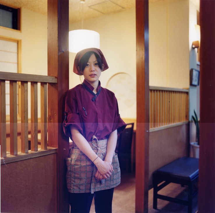 Yuka, a waitress at the Kuroshio restaurant, standing in a traditional Japanese interior space wearing purple clothing and a red headscarf.