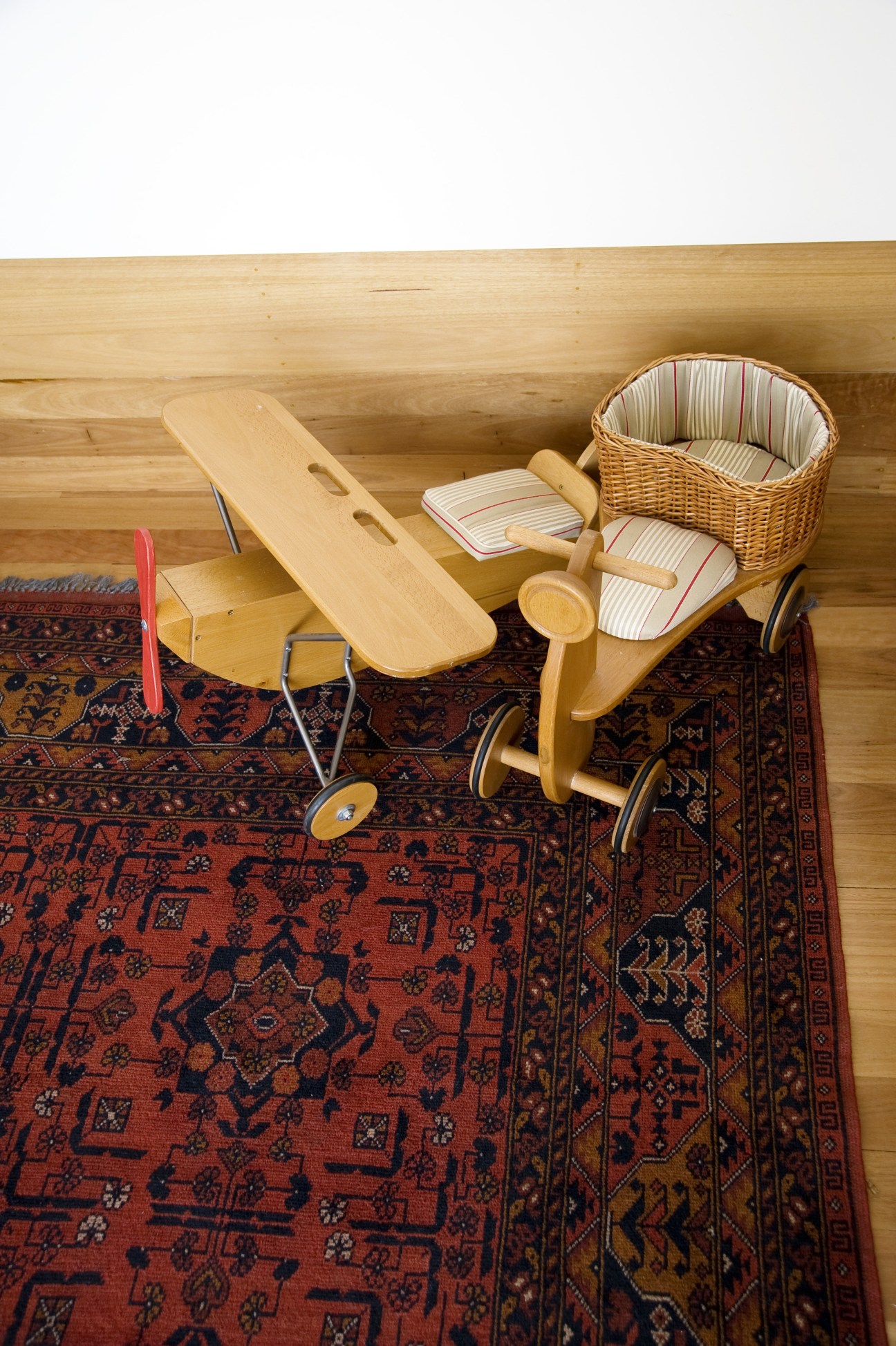 Wooden toy tricycles and a basket on a red patterned rug against wooden floors.
