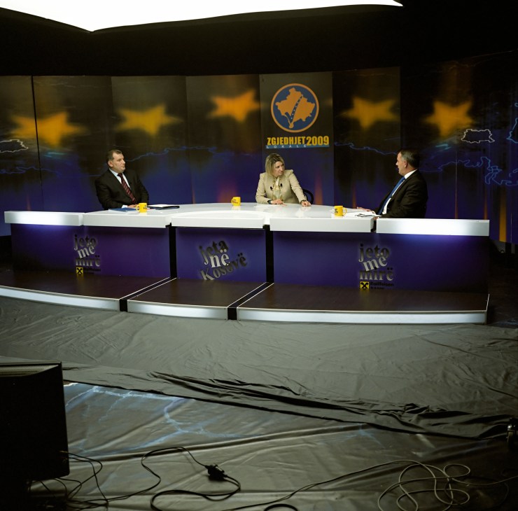Jeta Xharra interviewing a local politician on a television set with Kosovo branding and yellow stars in the background.