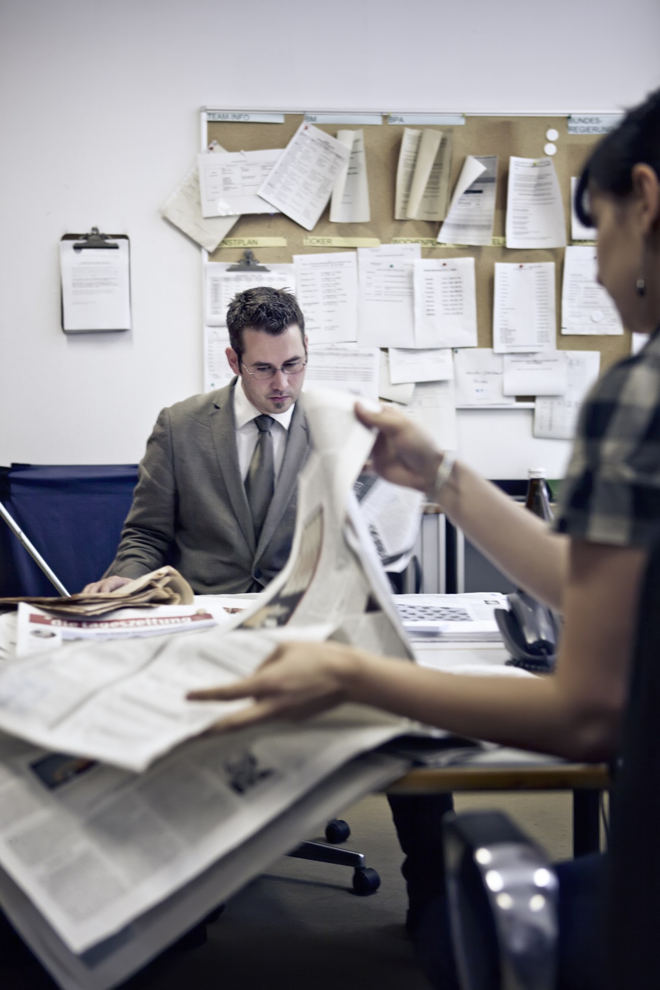 Two staff members work at a desk in the press department with documents and papers pinned to a board behind them.