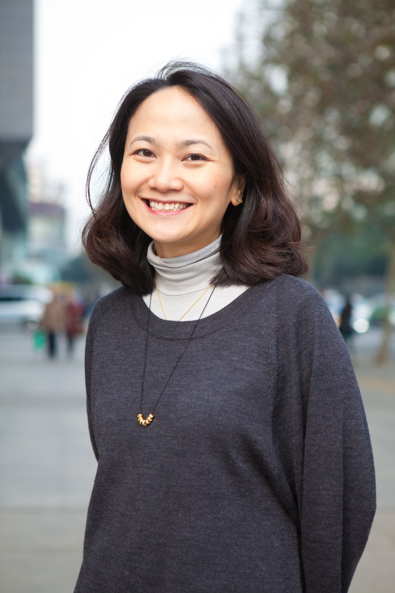 Woman smiling at camera wearing dark sweater and gold necklace with urban background