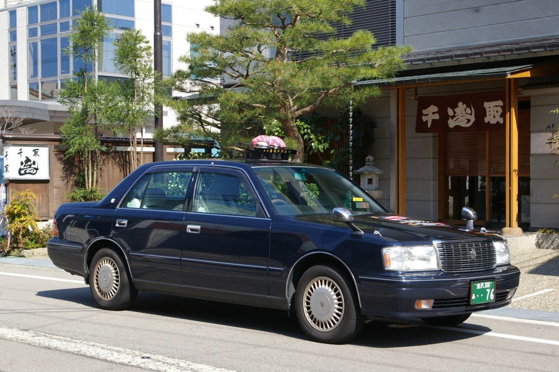 Black taxi cab with pink sushi decoration on roof parked outside traditional Japanese building in Kanazawa.