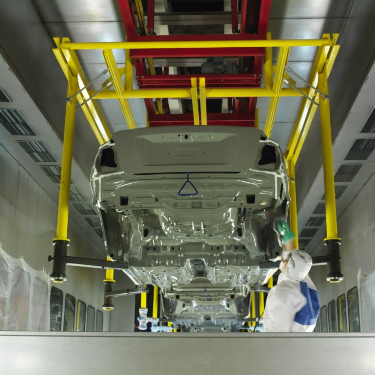 Worker in white coat and blue clothing operates yellow industrial machinery on a car assembly line at Avtotor plant.