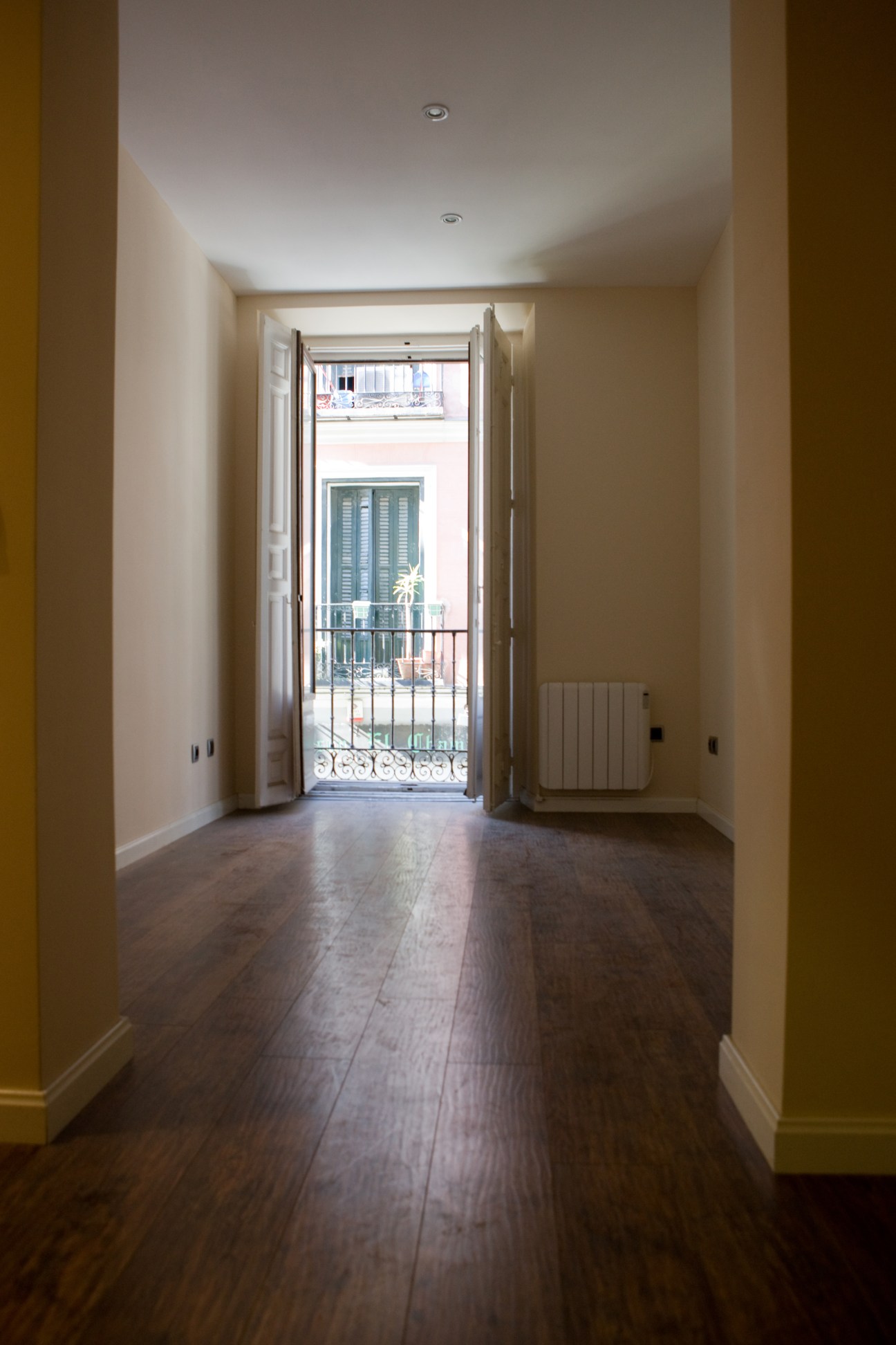 A bright hallway with wooden floors and cream walls opening to a balcony with ornate railing overlooking a Madrid street.