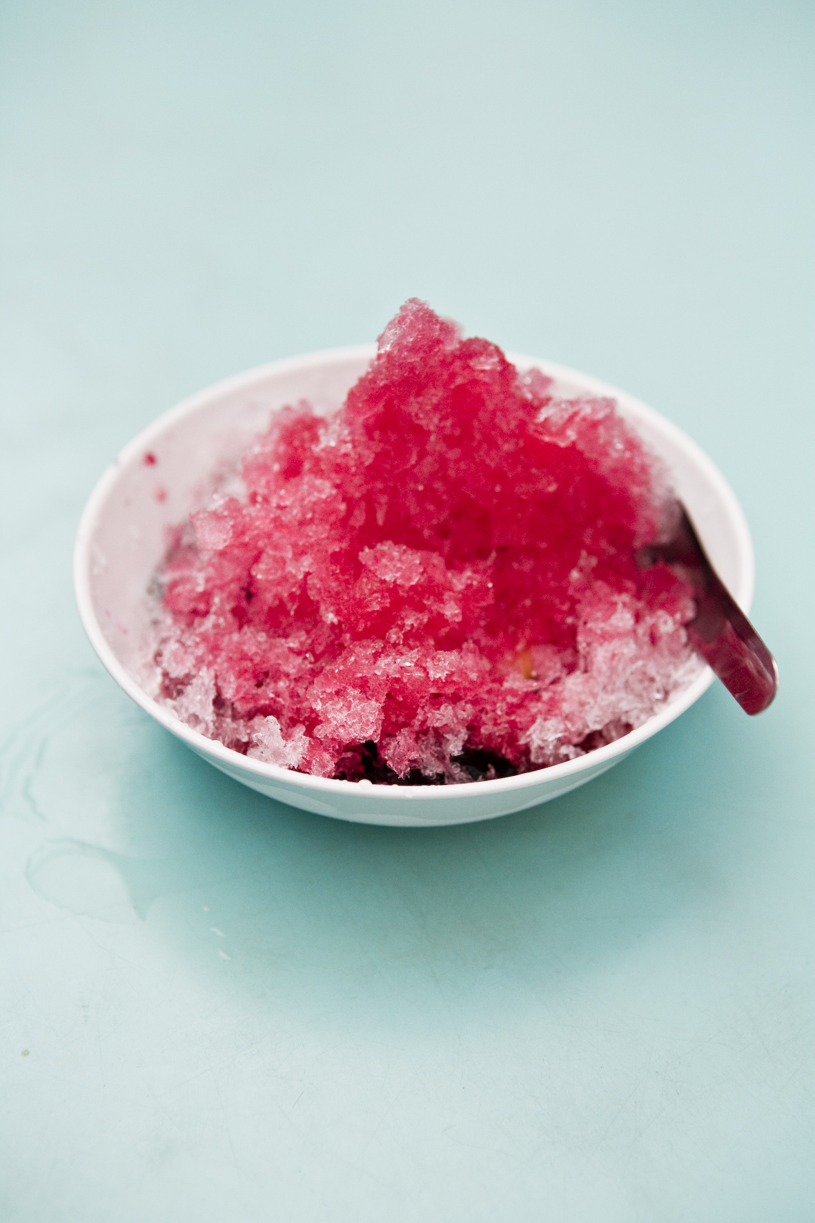 Pink shaved ice dessert in a white bowl with a wooden spoon on a light blue background.