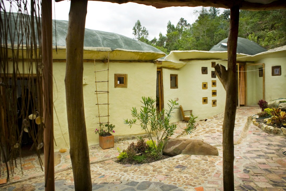 Yellow-walled ecological house with wooden pergola, stone courtyard and rolling green hills behind.