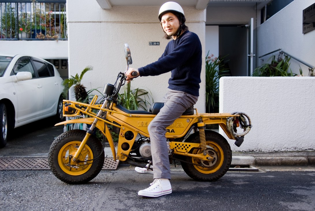 Designer Takuya Kawashima sits on a yellow Honda Motra scooter wearing a white helmet and dark sweater.