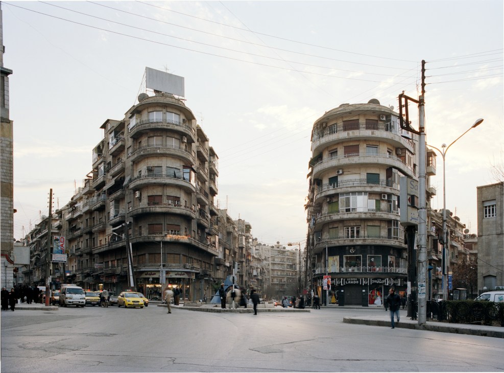 Modernist corner buildings with curved facades in Mother Teresa Square, Aleppo, with pedestrians and yellow taxis on the street.
