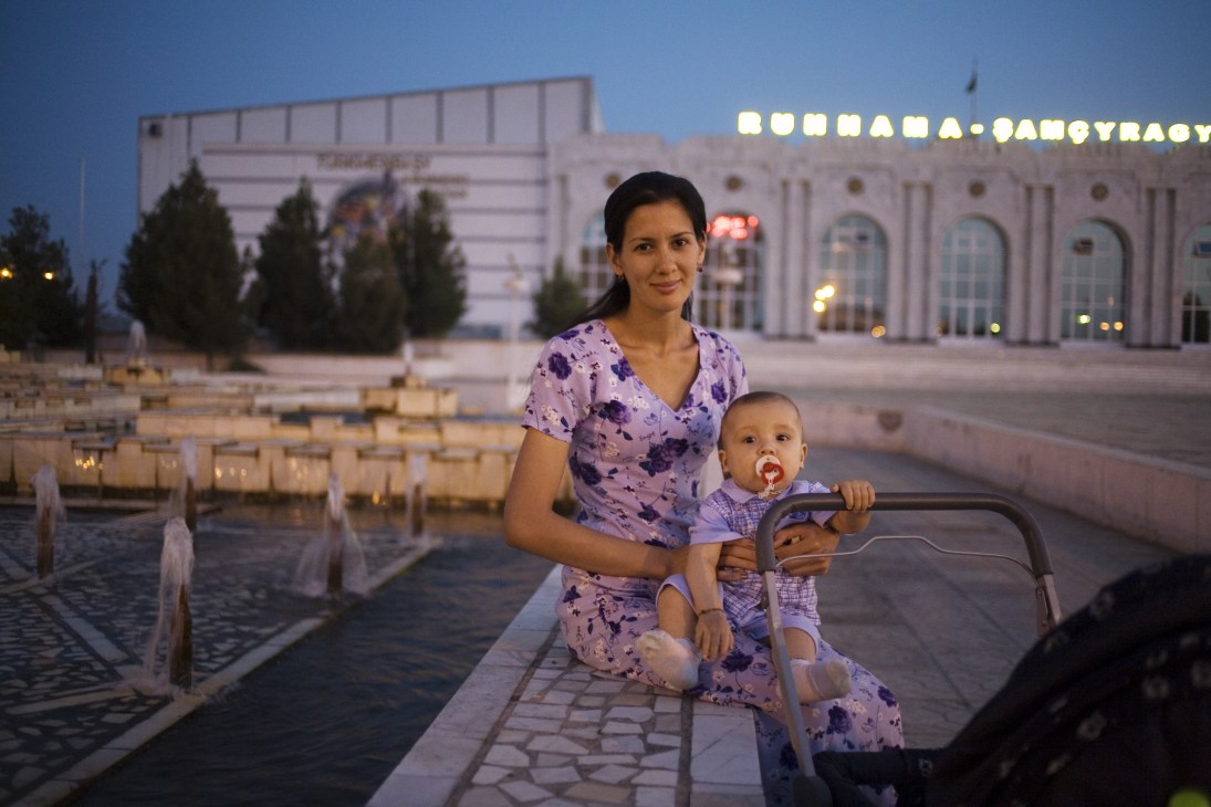A woman in a purple patterned dress holds a young child while sitting on a bench in a central Mary plaza, with an illuminated government building behind them at dusk.