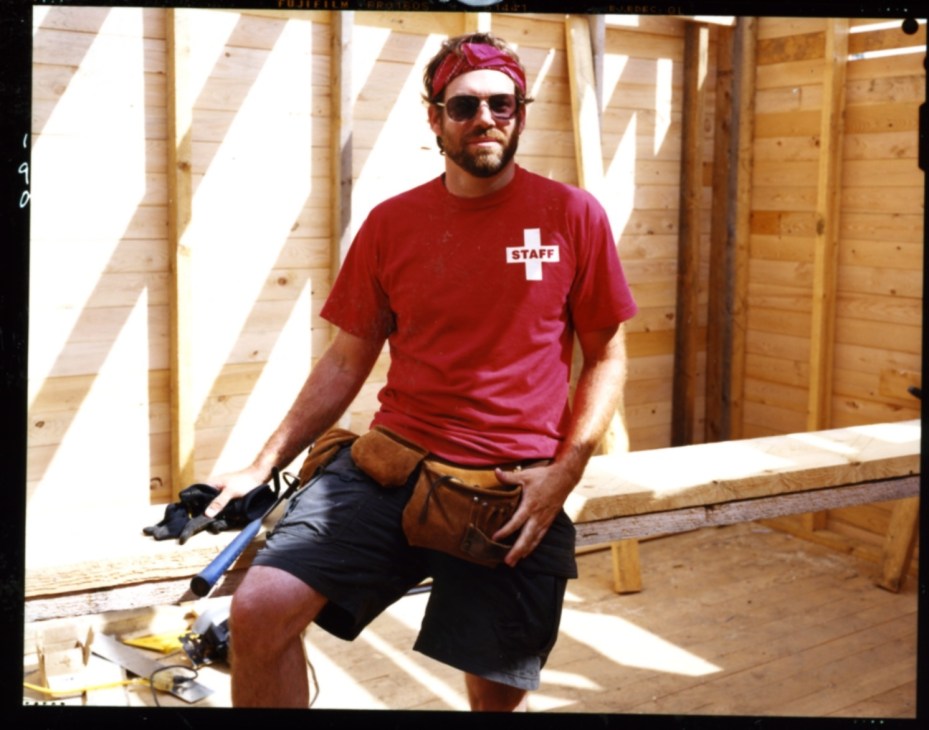 Architect Dale Rush sitting on wooden beams inside a building under construction, wearing a red staff t-shirt and tool belt.