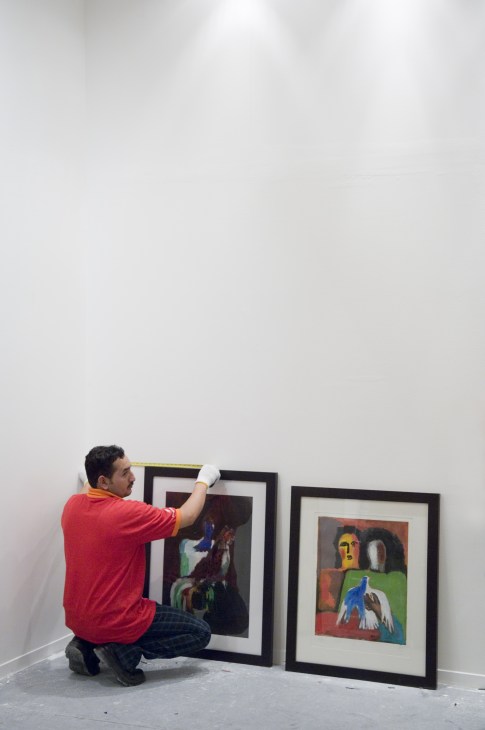 A gallerist in a red shirt measures and adjusts framed artworks during Art Dubai setup.