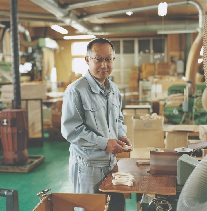 Fumihiko Koyama stands in his wooden toy workshop surrounded by machinery and wooden toy components.