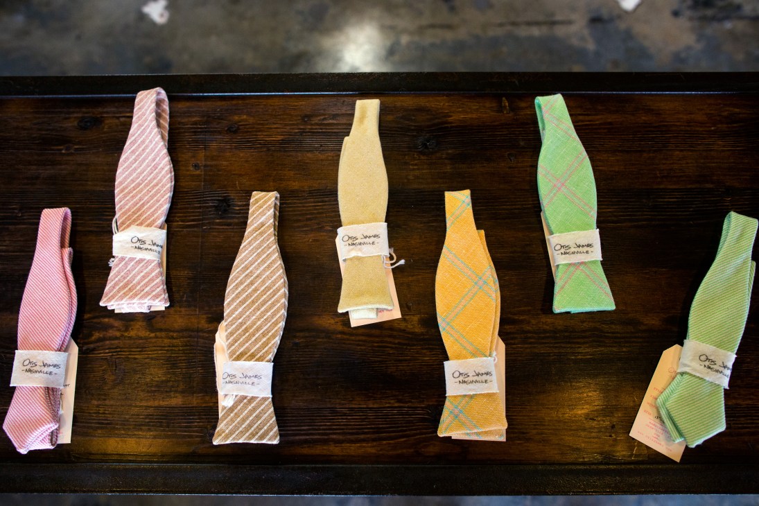 Colorful finished bow ties displayed on a dark wooden shelf at Otis James Studio.