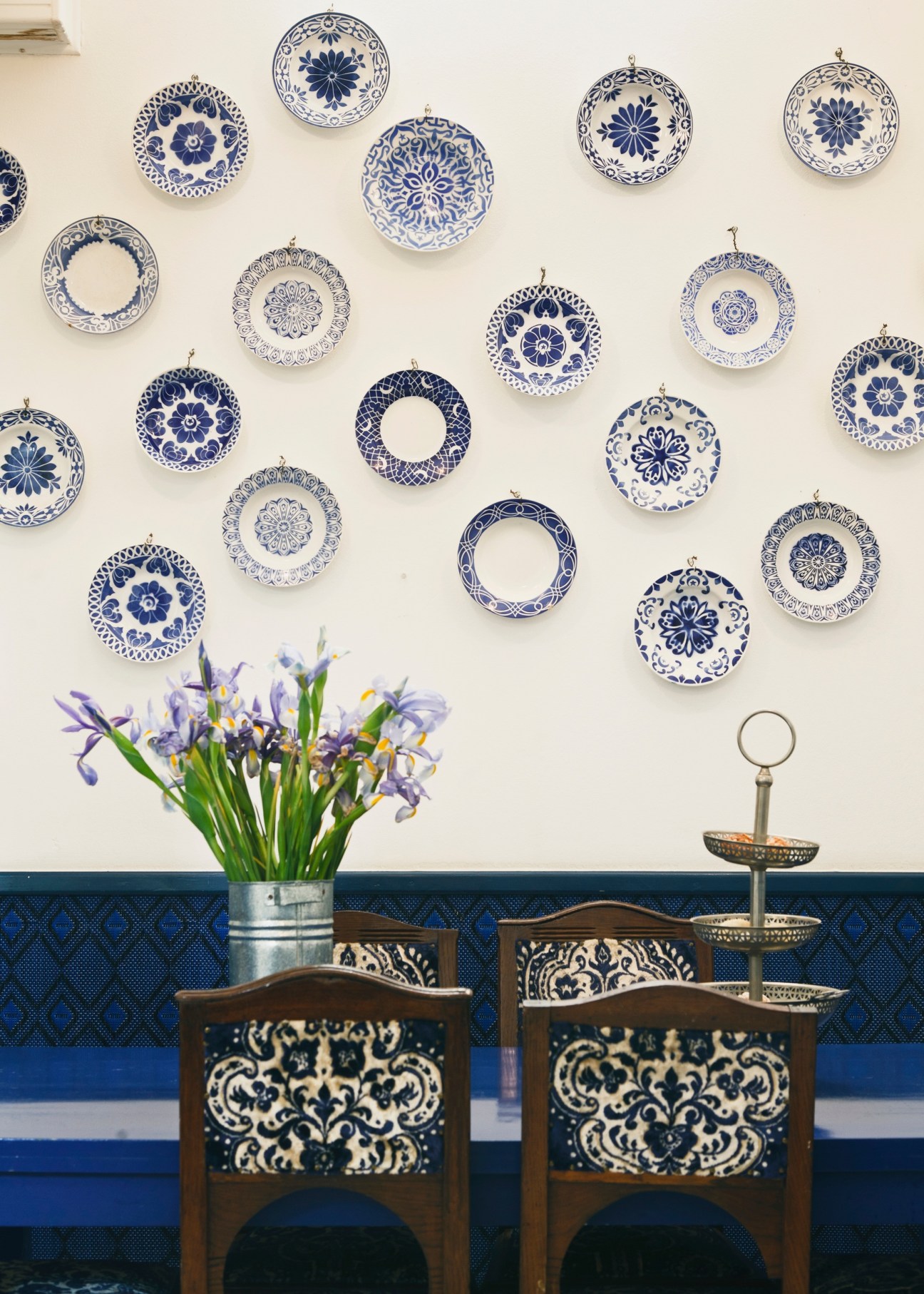 Blue and white decorative plates arranged on a cream wall above wooden chairs with patterned cushions and fresh irises in a vase.