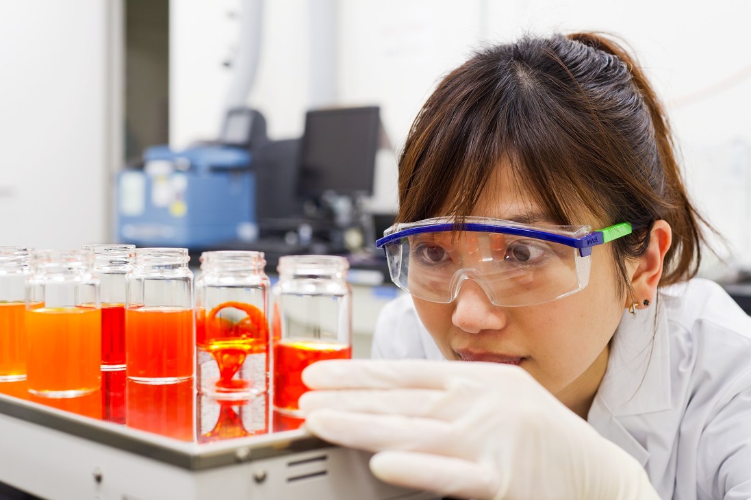 Researcher in safety goggles examining red liquid samples in laboratory containers.